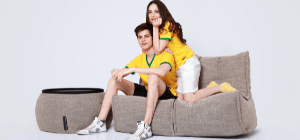 Australian Couple wearing yellow shirts sits on brown bean bag sofa bed with brown ottoman