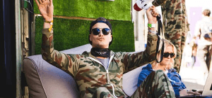 Australian guy wearing sunglasses holds megaphone wearing camouflage coverall sitting in brown cream nude bean bag sofa chair