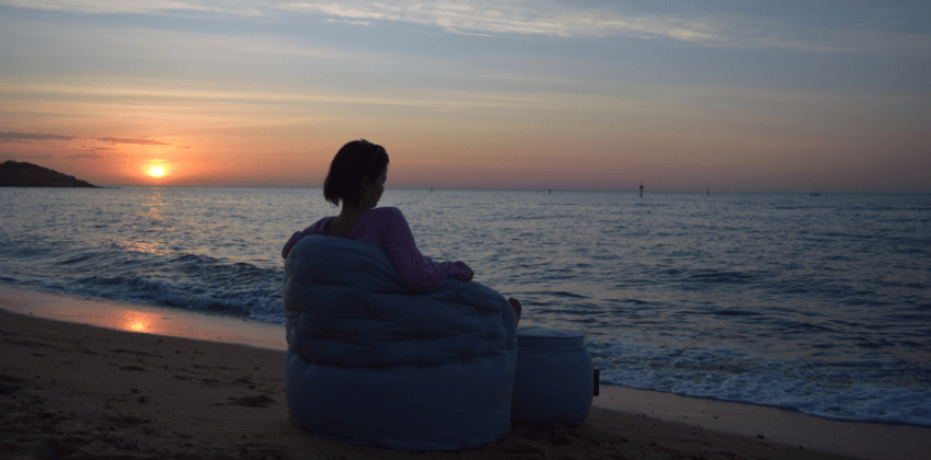 Woman sitting on a bean bag sofa chair with ottoman table facing the ocean with sunset background
